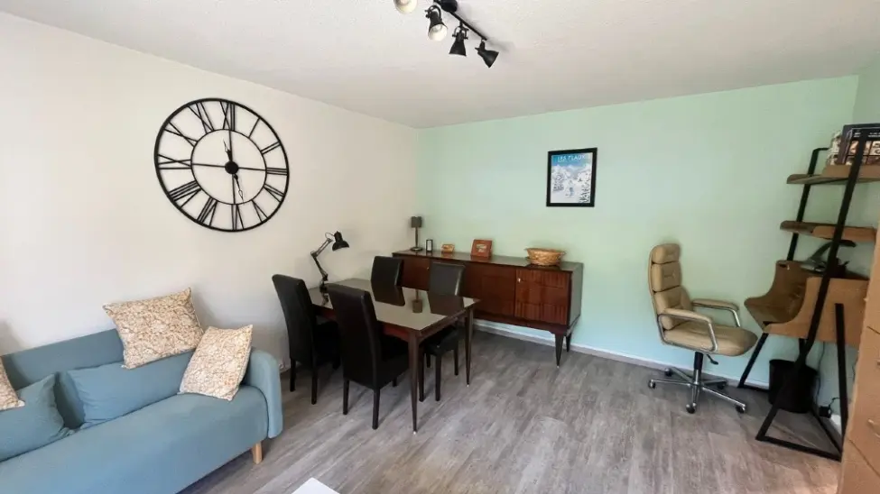 Un salon-salle à manger avec un sol en parquet flottant, un canapé turquoise sur la gauche, une table à manger rectangulaire au centre avec six chaises, un buffet en bois sombre le long du mur et un bureau avec une chaise de bureau sur la droite.