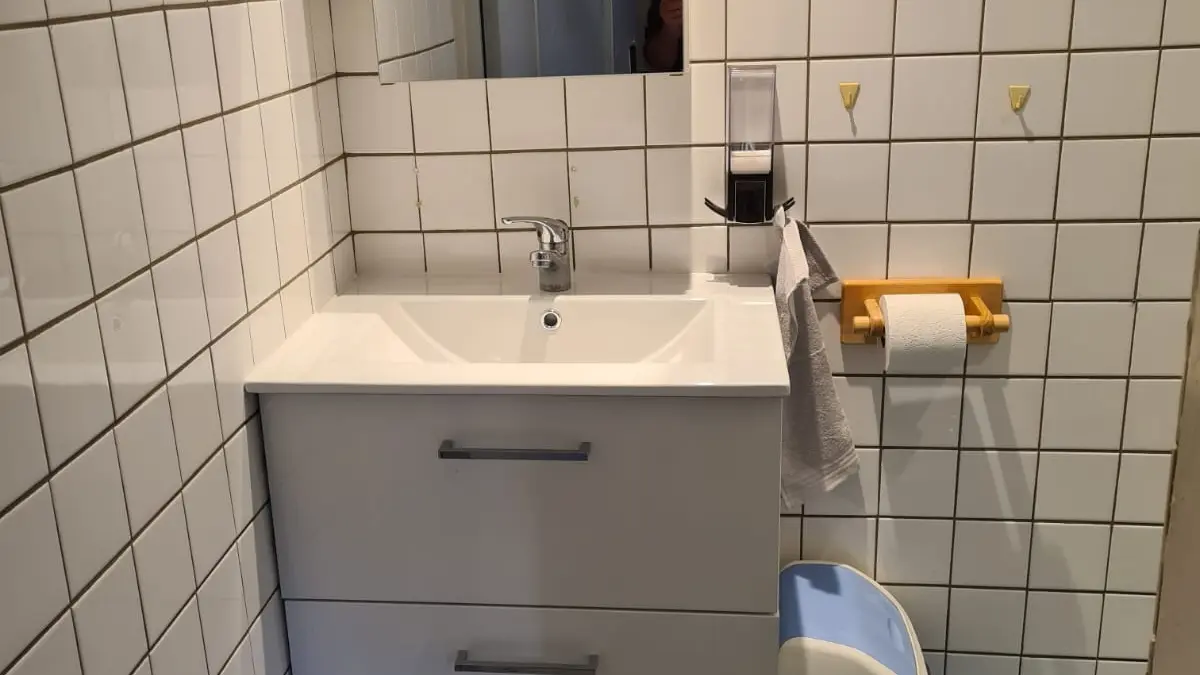 Cette salle d'eau est équipée d'un lavabo, d'un miroir, d'un distributeur de savon, d'un porte-serviette et d'une poubelle. Les murs et le sol sont carrelés.