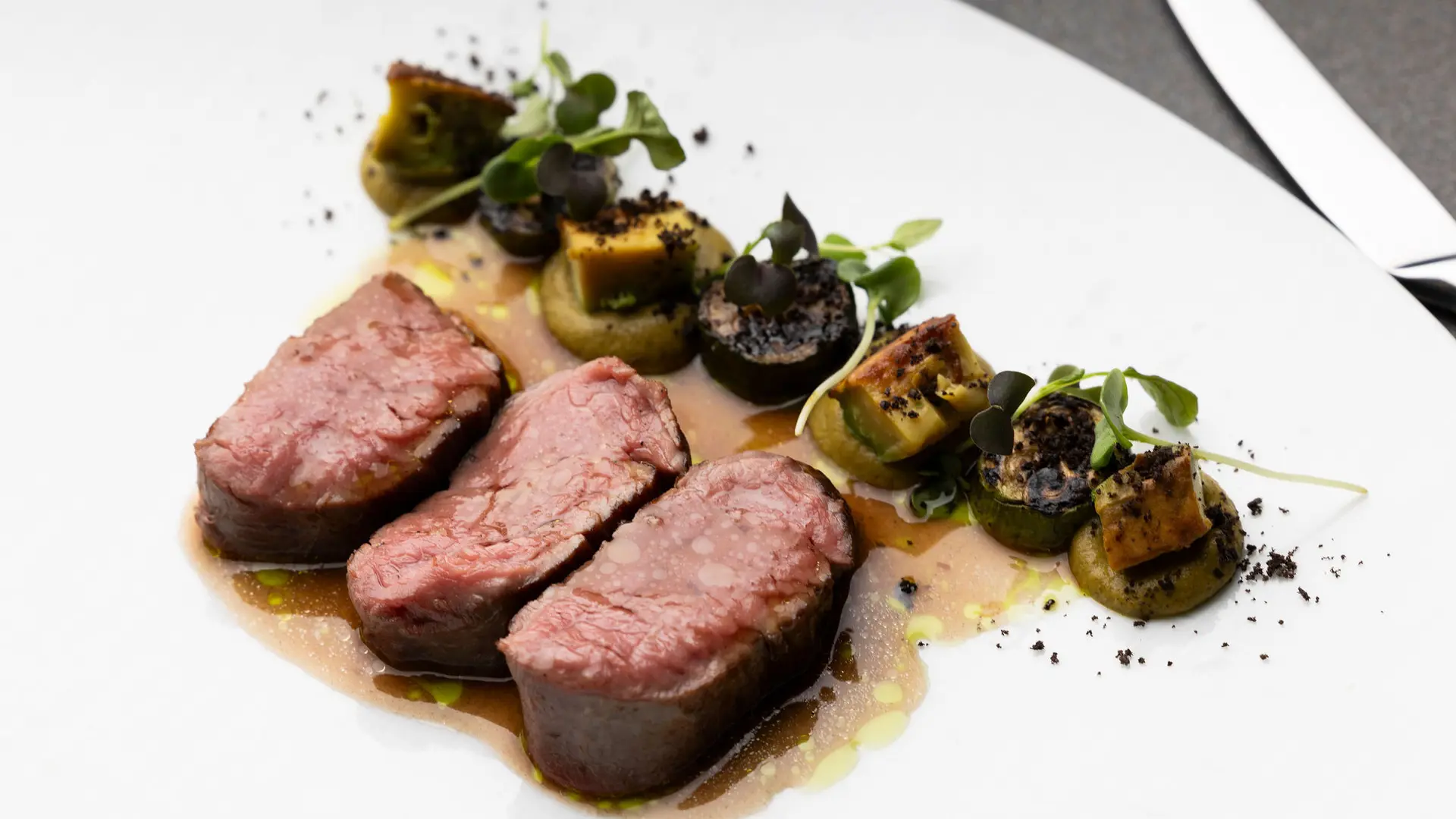 Trois médaillons de viande rouge cuite reposent sur une sauce légère, accompagnés d'une rangée de rondelles de légumes, de pousses vertes et de quelques miettes foncées.Trois médaillons de viande rouge cuite reposent sur une sauce légère, accompagnés d'une rangée de rondelles de légumes, de pousses vertes et de quelques miettes foncées.