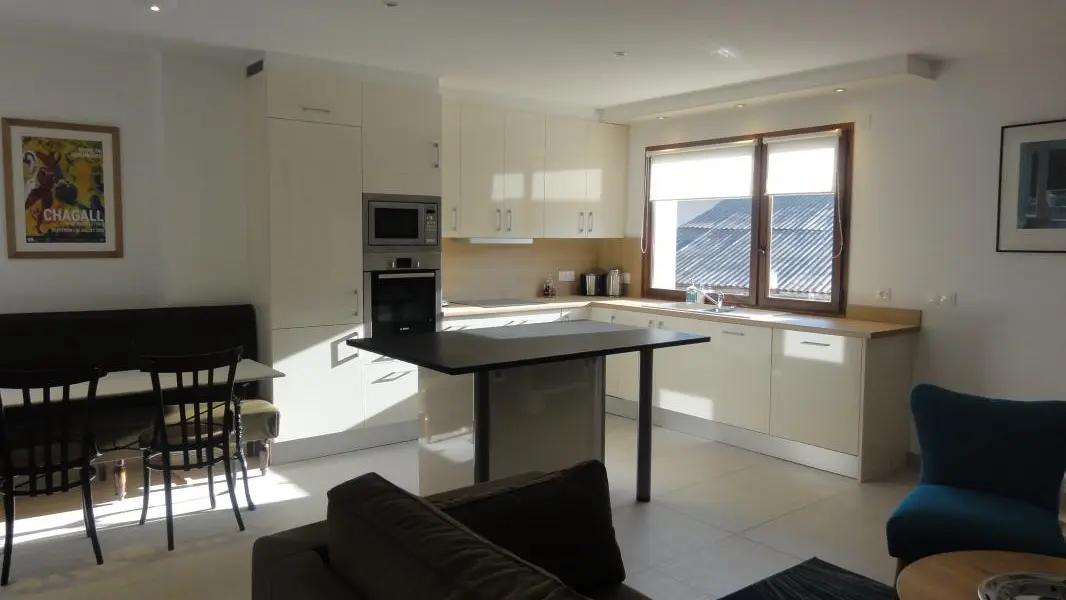 Ambiance d'un matin d'été avec vue sur la cuisine équipée avec son îlot central et l'espace repas à gauche (table en marbre, banquette et chaises bistrots).