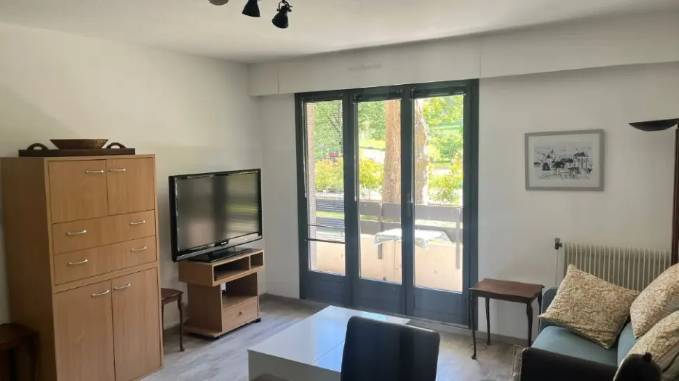 Un salon avec des murs blancs, un meuble de rangement en bois clair, une télévision, un canapé turquoise, une table basse blanche, et une large baie vitrée donnant sur l'extérieur.