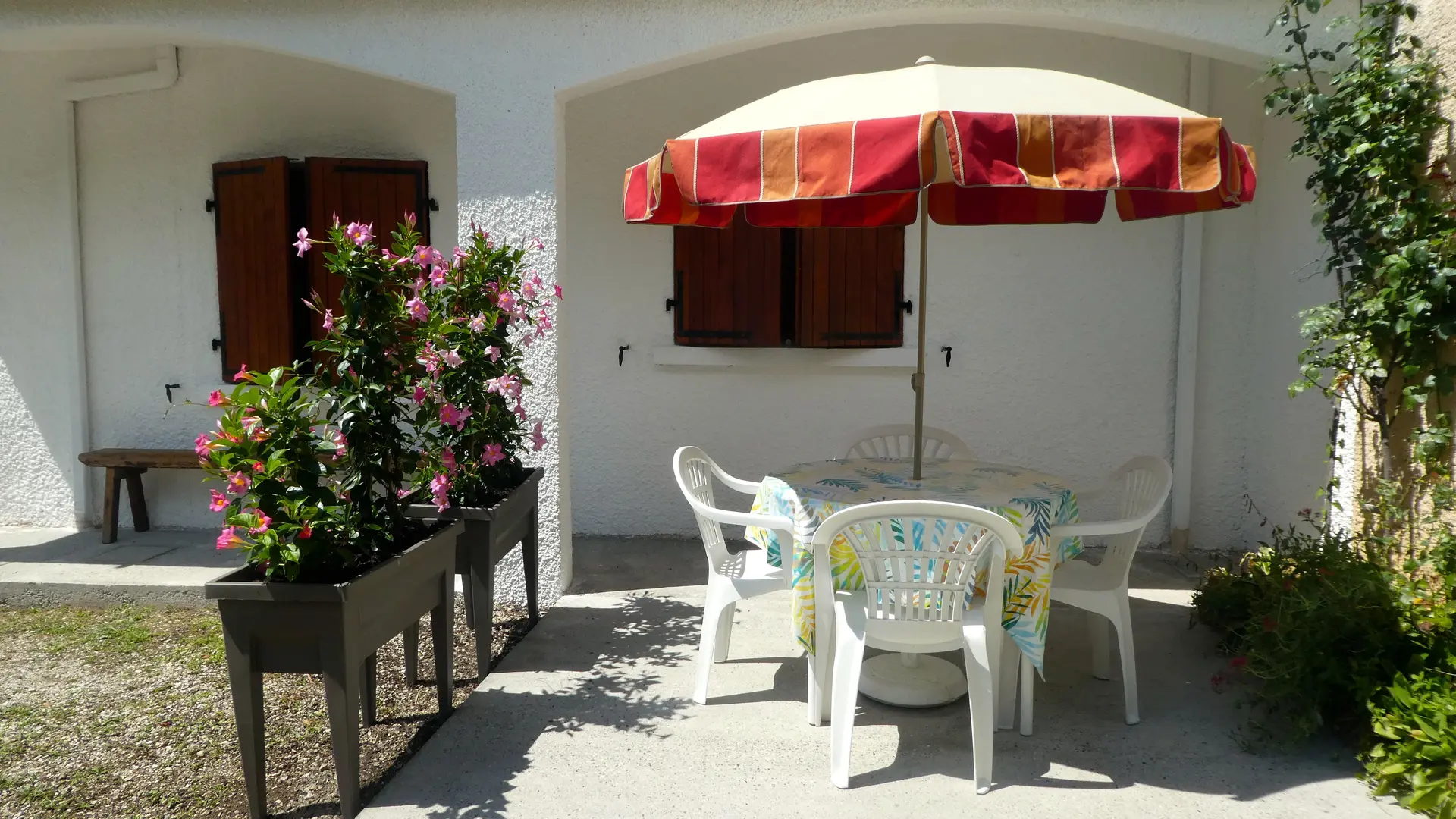 La terrasse extérieure avec une petite table de jardin entourée de chaises blanches, un parasol rouge et blanc offrant de l'ombre.