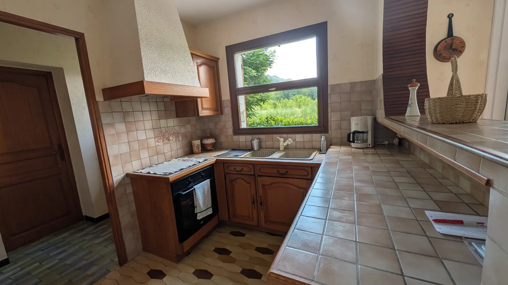Une cuisine équipée en U avec un plan de travail et une crédence en carreaux, un sol en carrelage, un meuble bas pour un four à gauche et une fenêtre centrale qui donne sur l'extérieur.