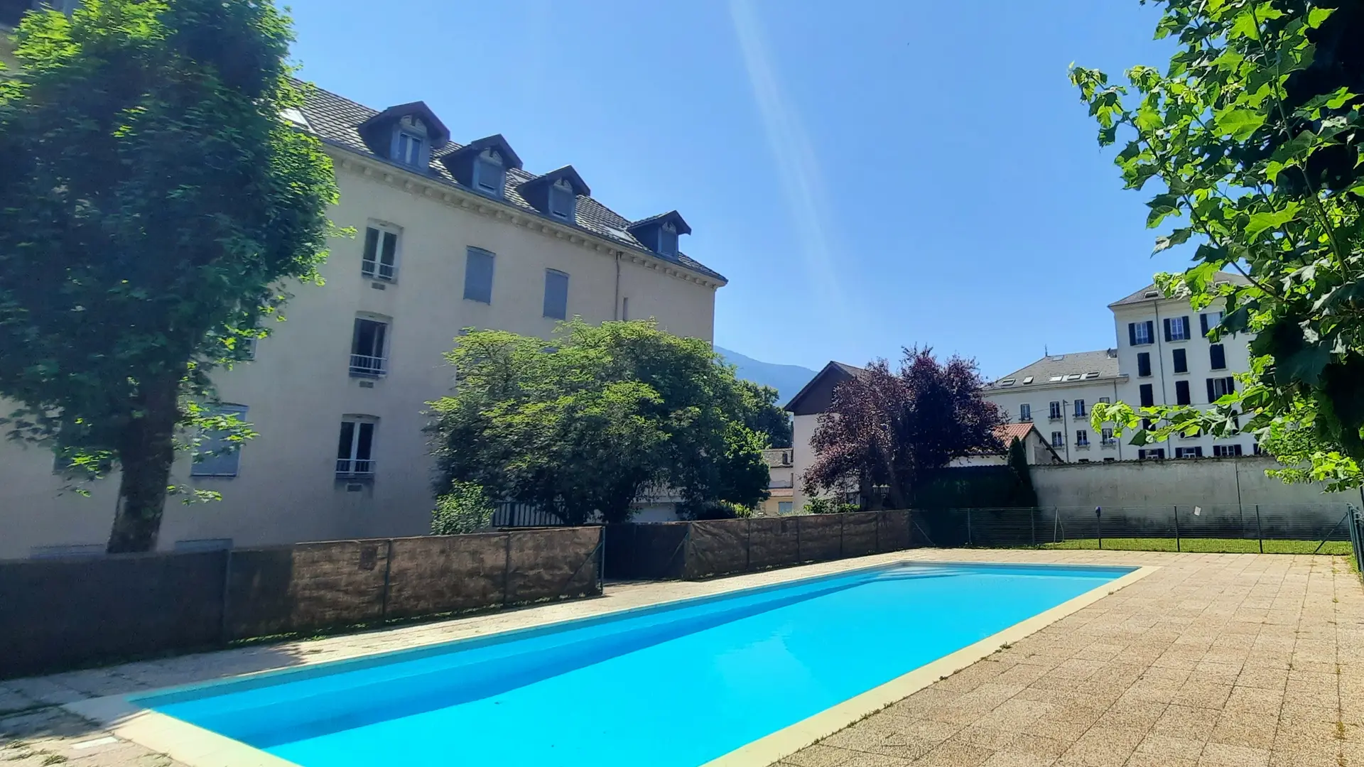 Cette vue donne sur une piscine rectangulaire, entourée d'une pelouse et bordée d'un immeuble.