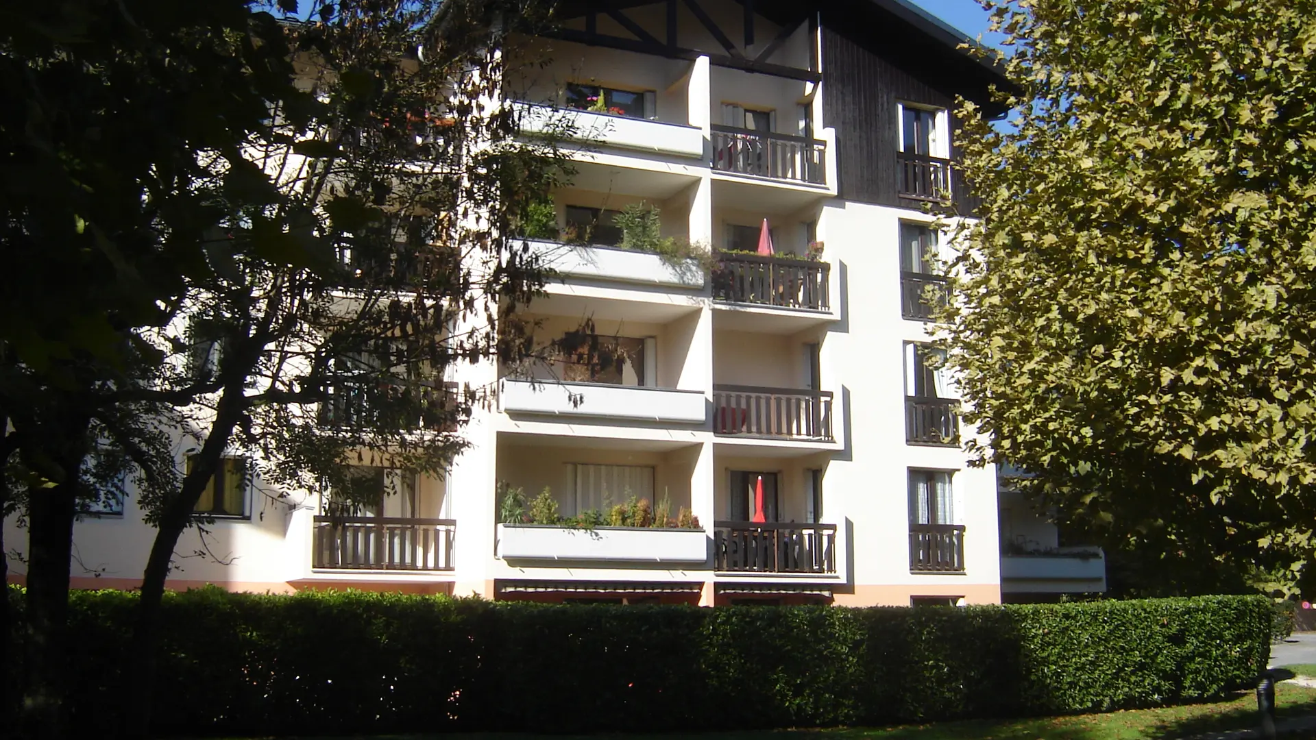 L'Orée du Parc est un bâtiment de plusieurs étages, avec un toit pentu qui rappelle un chalet. Les murs sont principalement blancs et en bois sur certaines parties. Le bâtiment est entouré d'arbres qui créent un environnement frais et ombragé.