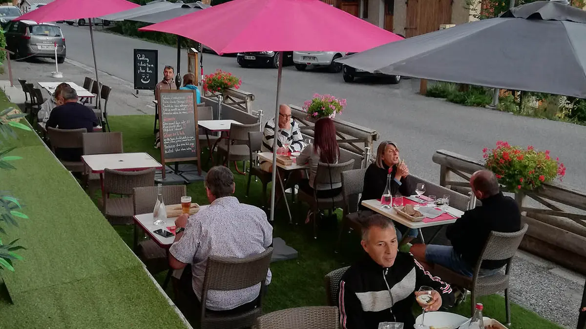 Terrasse extérieure avec sol en faux gazon, tables et chaises, parasols, grand-rue du village