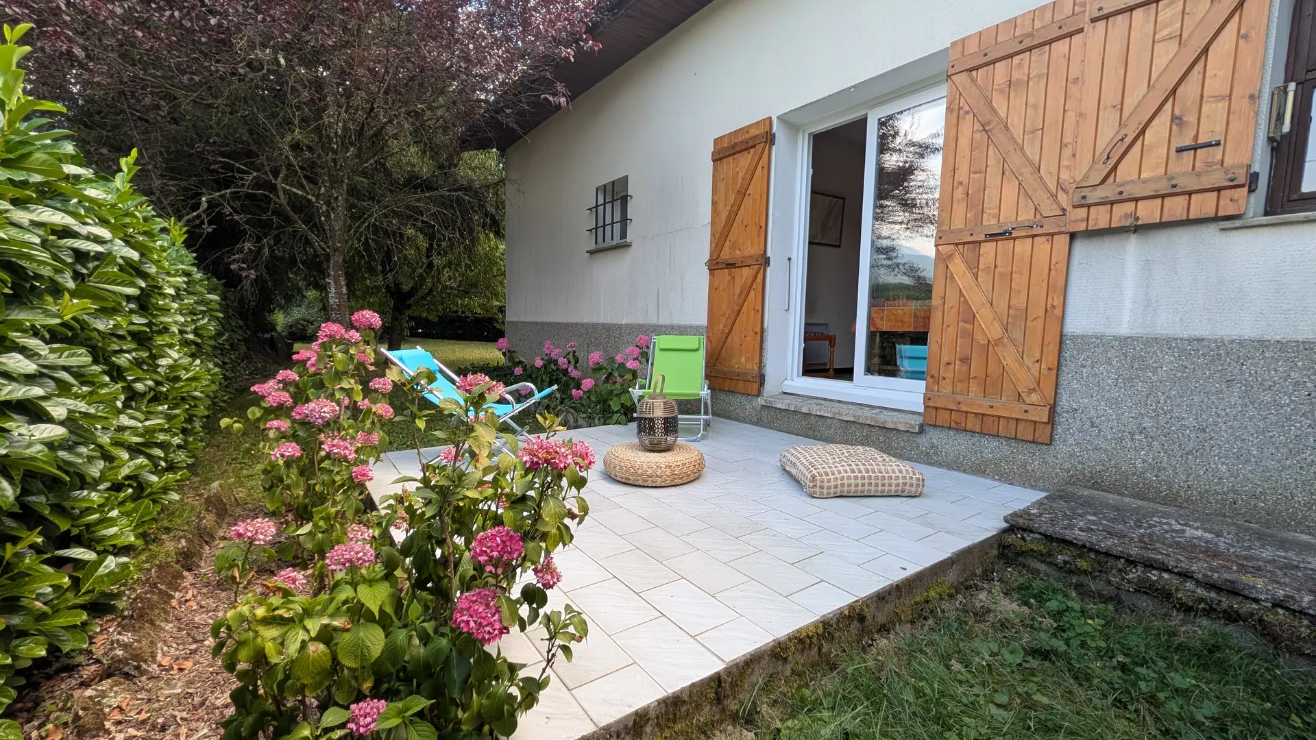 Cette terrasse carrelée, bordée de verdure et de parterres de fleurs, s'ouvre sur l'intérieur grâce à une porte-fenêtre.