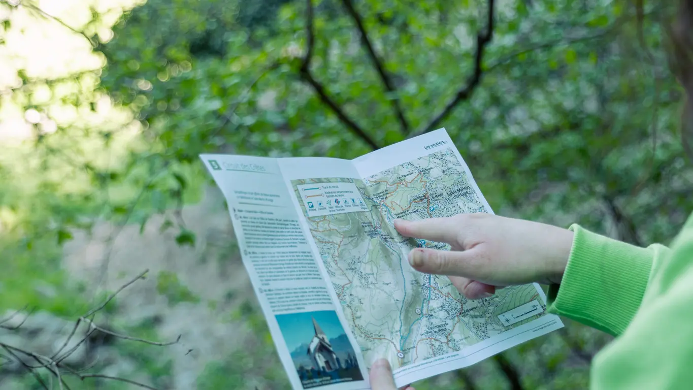Une personne portant un vêtement vert tient une carte en plein air, au bord d’un chemin ou d’un pont en bois, entourée de végétation dense et surplombant un ruisseau rocheux.