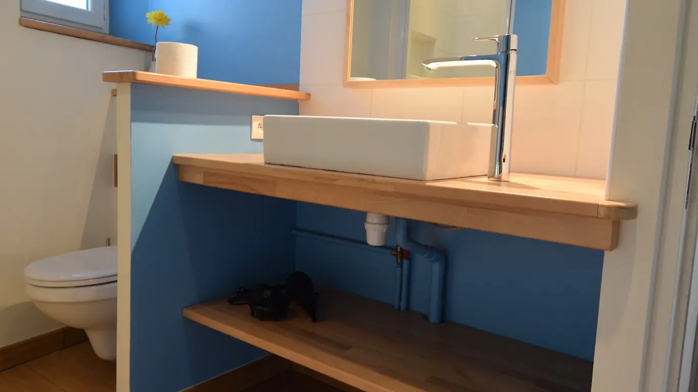 La salle de bains présente un design épuré avec un mur bleu vif contrastant avec le meuble en bois naturel du lavabo. Le grand miroir encastré dans le mur bleu apporte de la luminosité à la pièce. Le plan de travail du lavabo est en bois massif, tandis que les étagères inférieures offrent un espace de rangement supplémentaire.