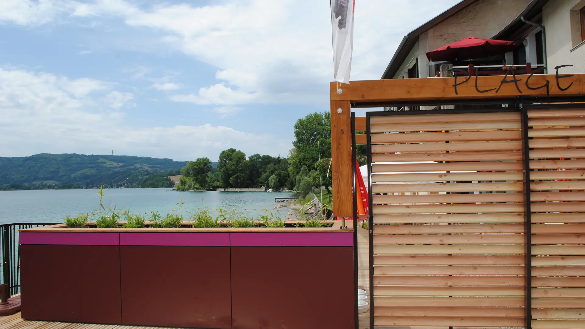 vue directe sur l'entrée de la plage depuis la terrasse du snack. Petit muret et portail en bois fermé, vue du lac en arriere plan.