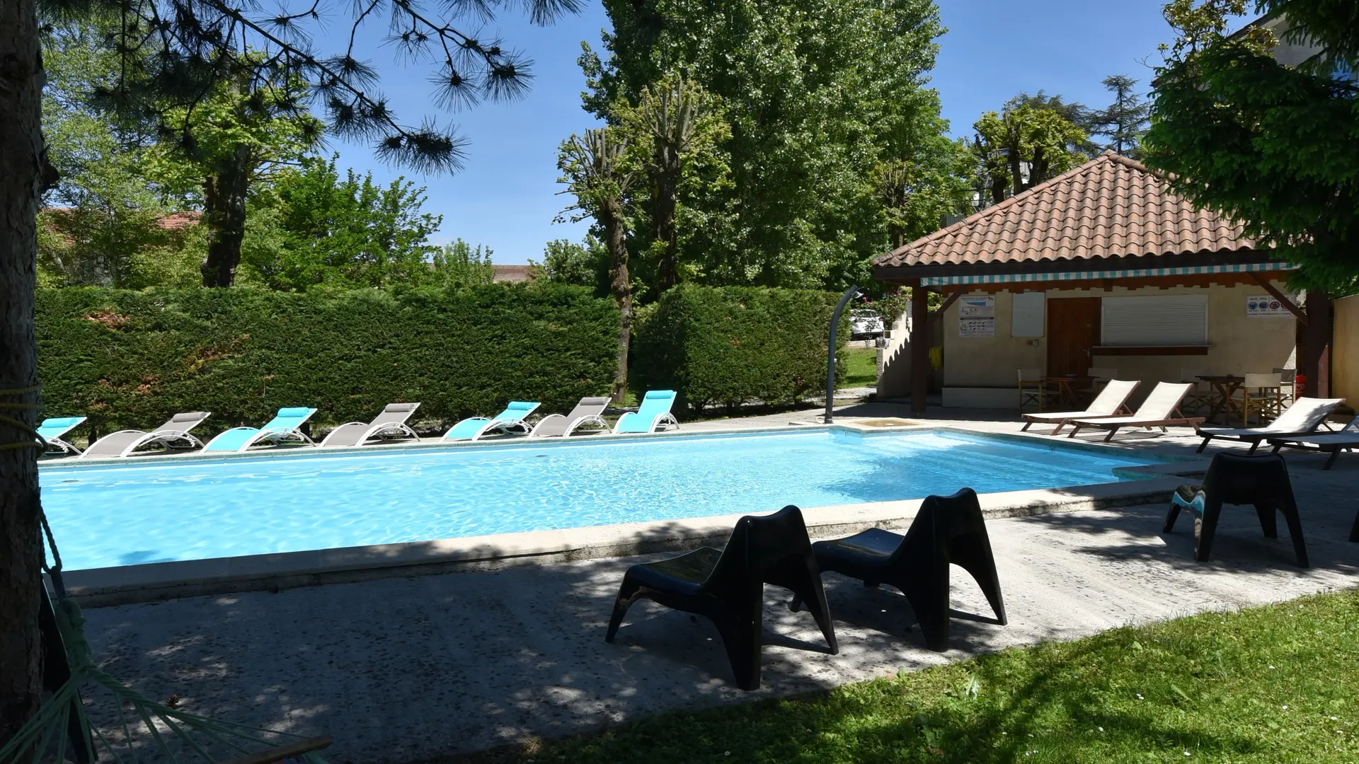 Espace piscine entouré d’arbres et comprenant des transats, des sièges, un hamac en premier plan, le tout installé devant le local technique