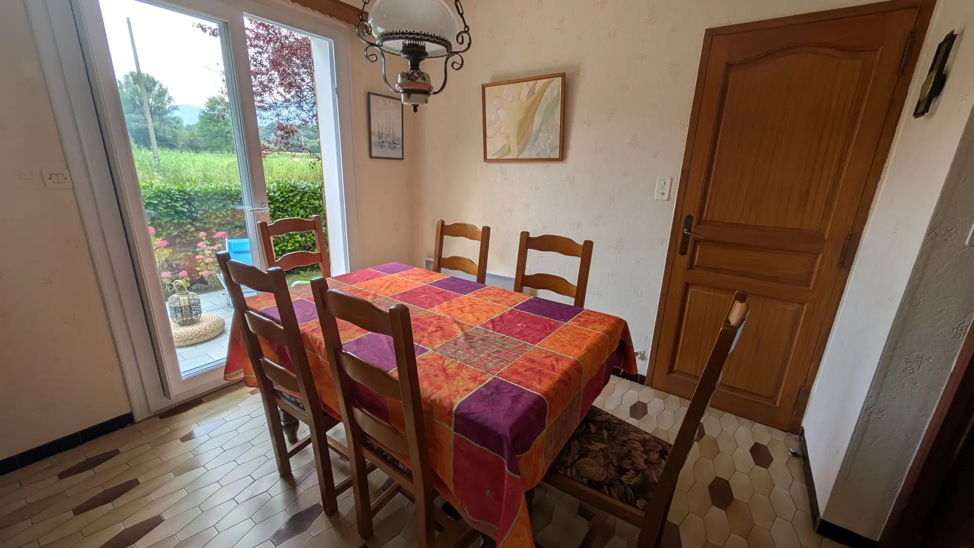 La pièce comporte une table à manger en bois et des chaises, située à côté d'une porte-fenêtre donnant sur la terrasse et d'une porte en bois donnant accès aux chambres.
