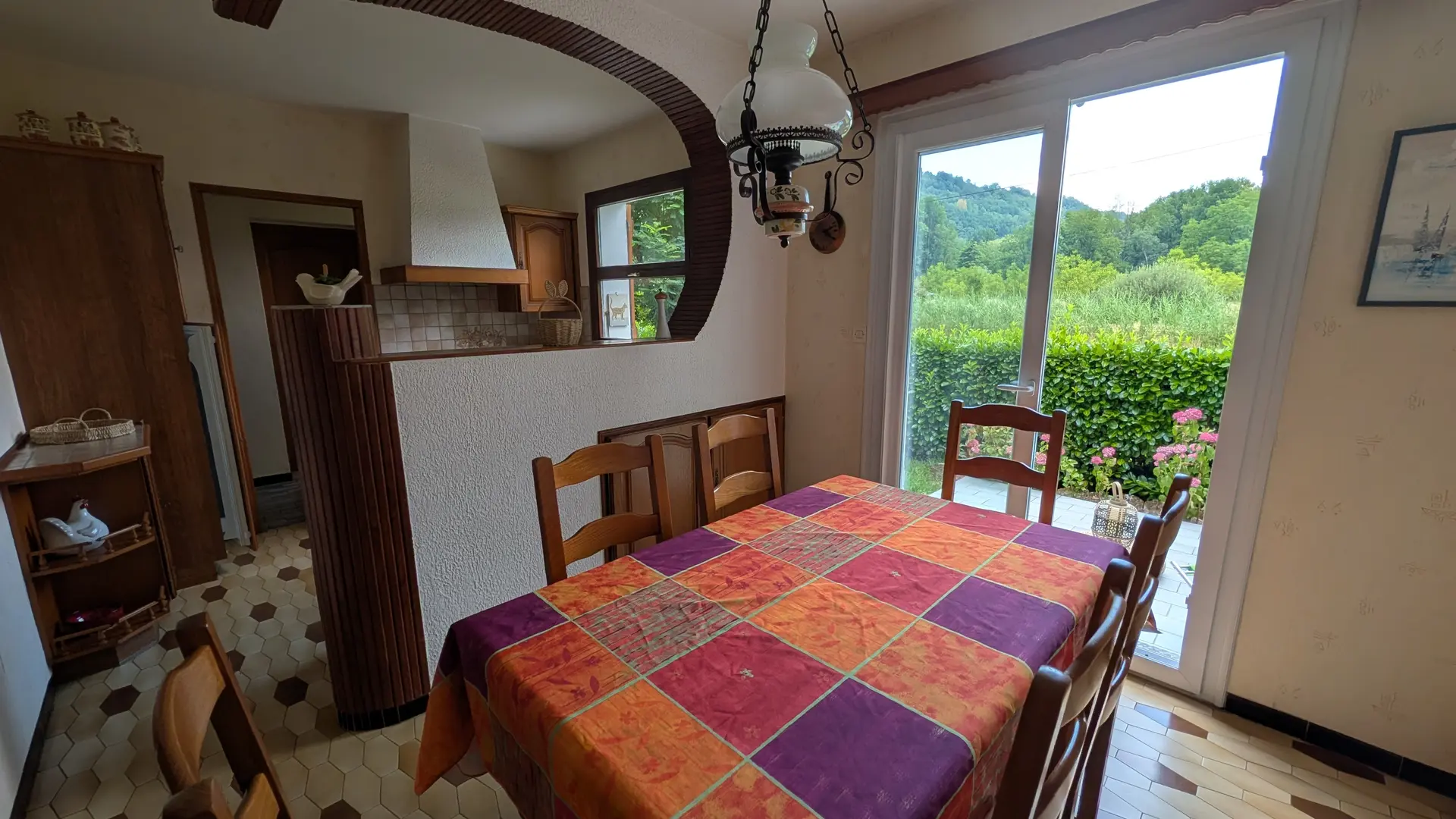 Une salle à manger meublée d'une table et de chaises en bois, qui donne sur une terrasse par une porte-fenêtre et s'ouvre sur une cuisine à l'arrière.
