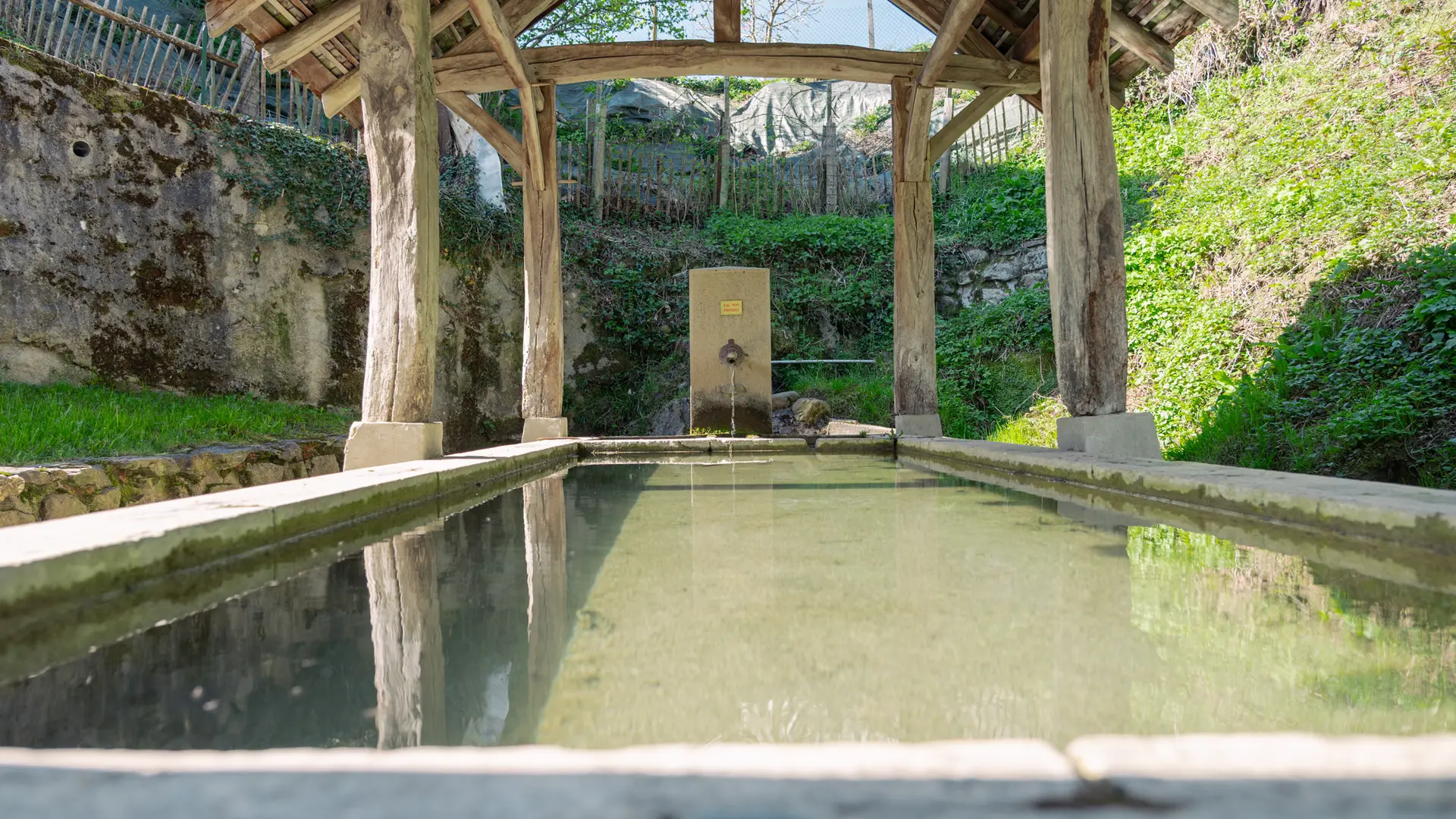 Il s'agit d'un lavoir rempli d'eau calme reflétant un cadre architectural en bois avec des poutres et des piliers, flanqué de murs en pierre recouverts de végétation, sous un ciel partiellement visible.
