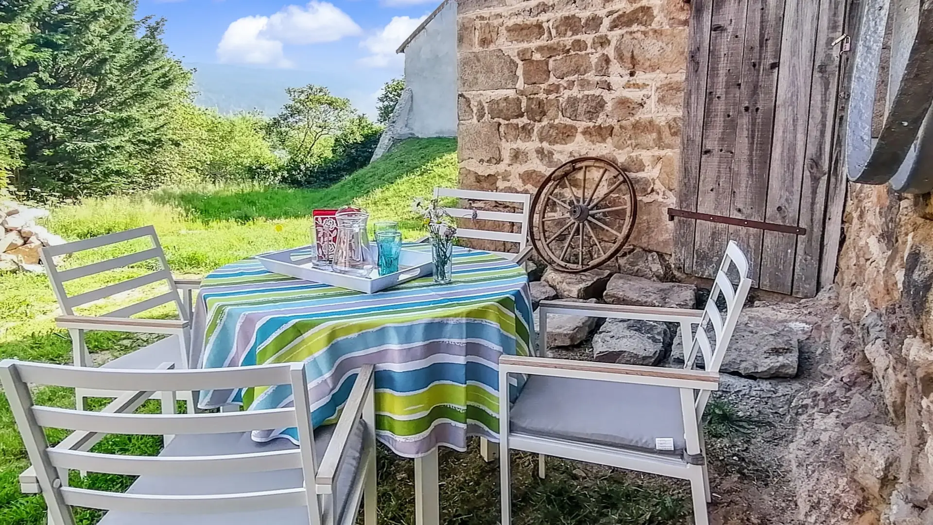 Une terrasse herbeuse avec une table et chaises et des boissons. A droite, on voit une roue à bandage accolée sur un mur en pierre, à côté une vielle porte en bois ainsi que des vieux objets