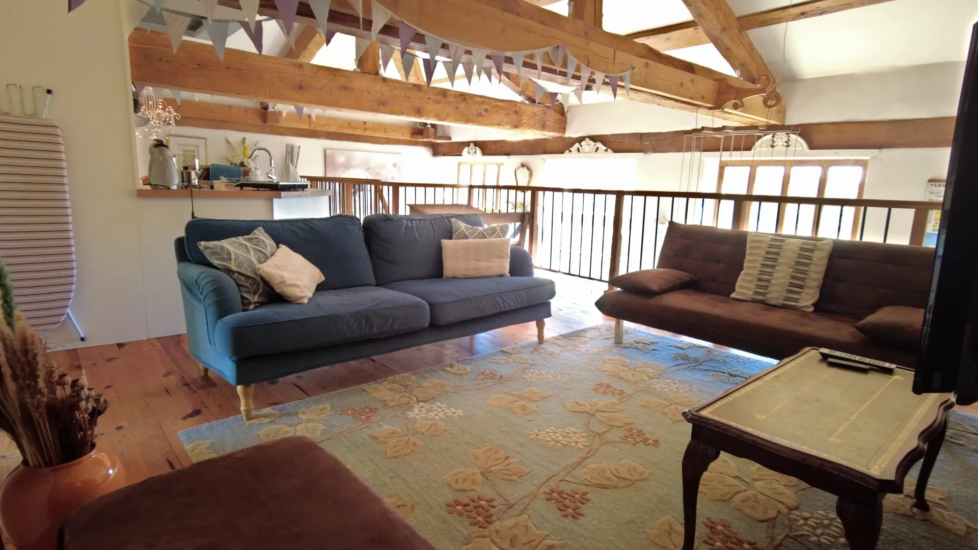 Nous sommes dans le salon de la mezzanine. Il comprend deux canapés : un bleu avec des coussins et un marron. Le plancher est recouvert d'un tapis. Une table basse se trouve en dessous de la télé. Du salon, on a une vue de la mezzanine avec sa rambarde.