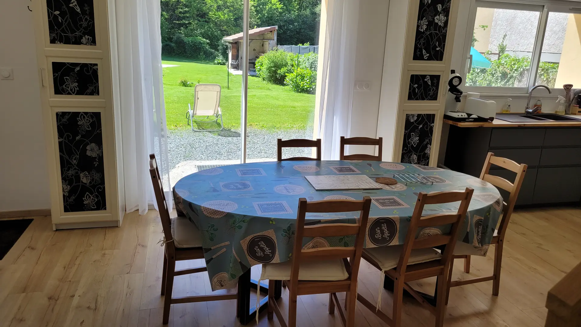 Cette pièce lumineuse donne sur un jardin par une grande baie vitrée. Au centre, une table ovale en bois, recouverte d'une nappe, est entourée de six chaises assorties. Le sol est en parquet clair et des étagères en bois sont disposées le long des murs.