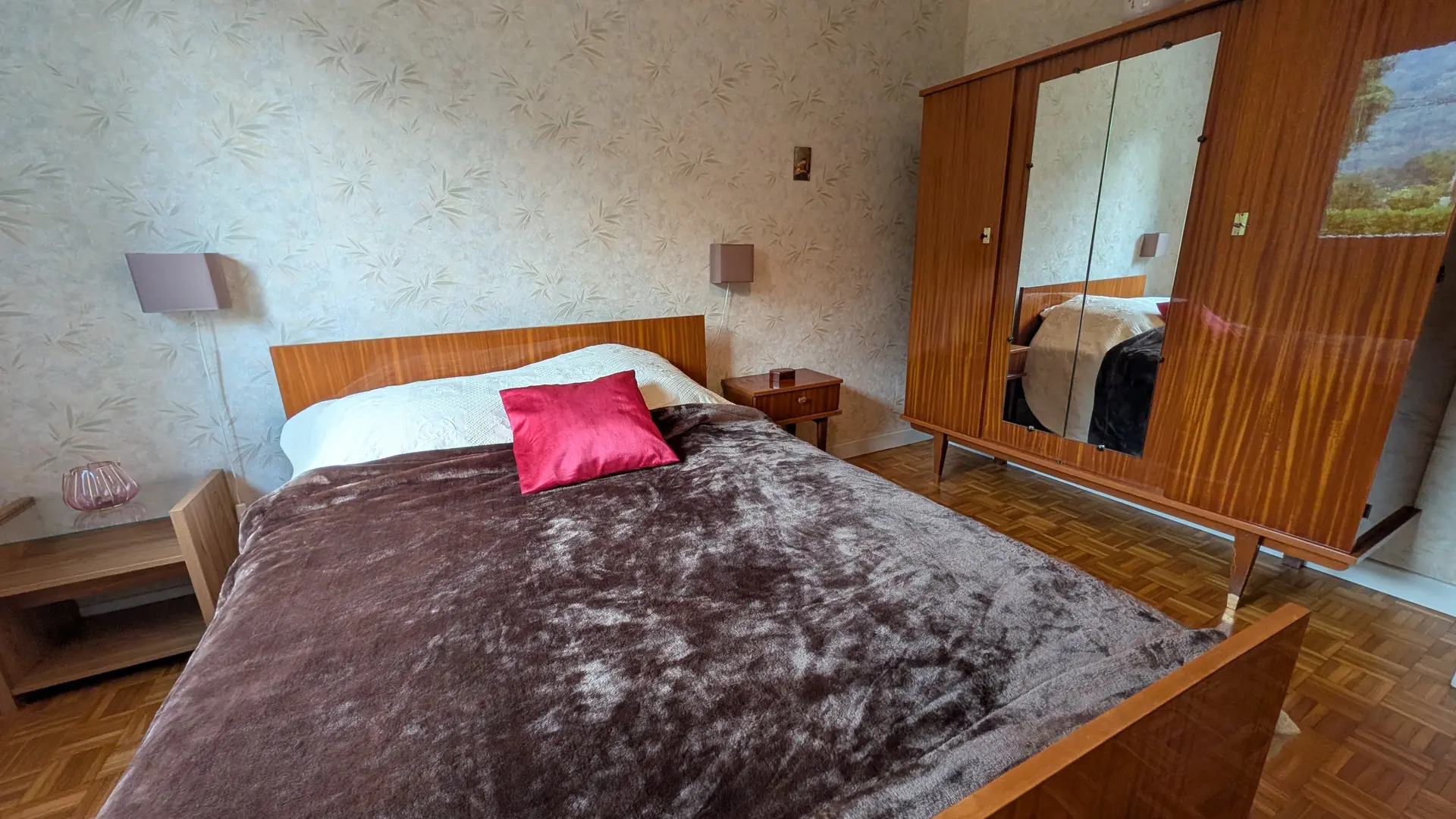 Une chambre à coucher présente un lit en bois, placé à côté d'une grande armoire en bois munie de miroirs sur le mur de droite.