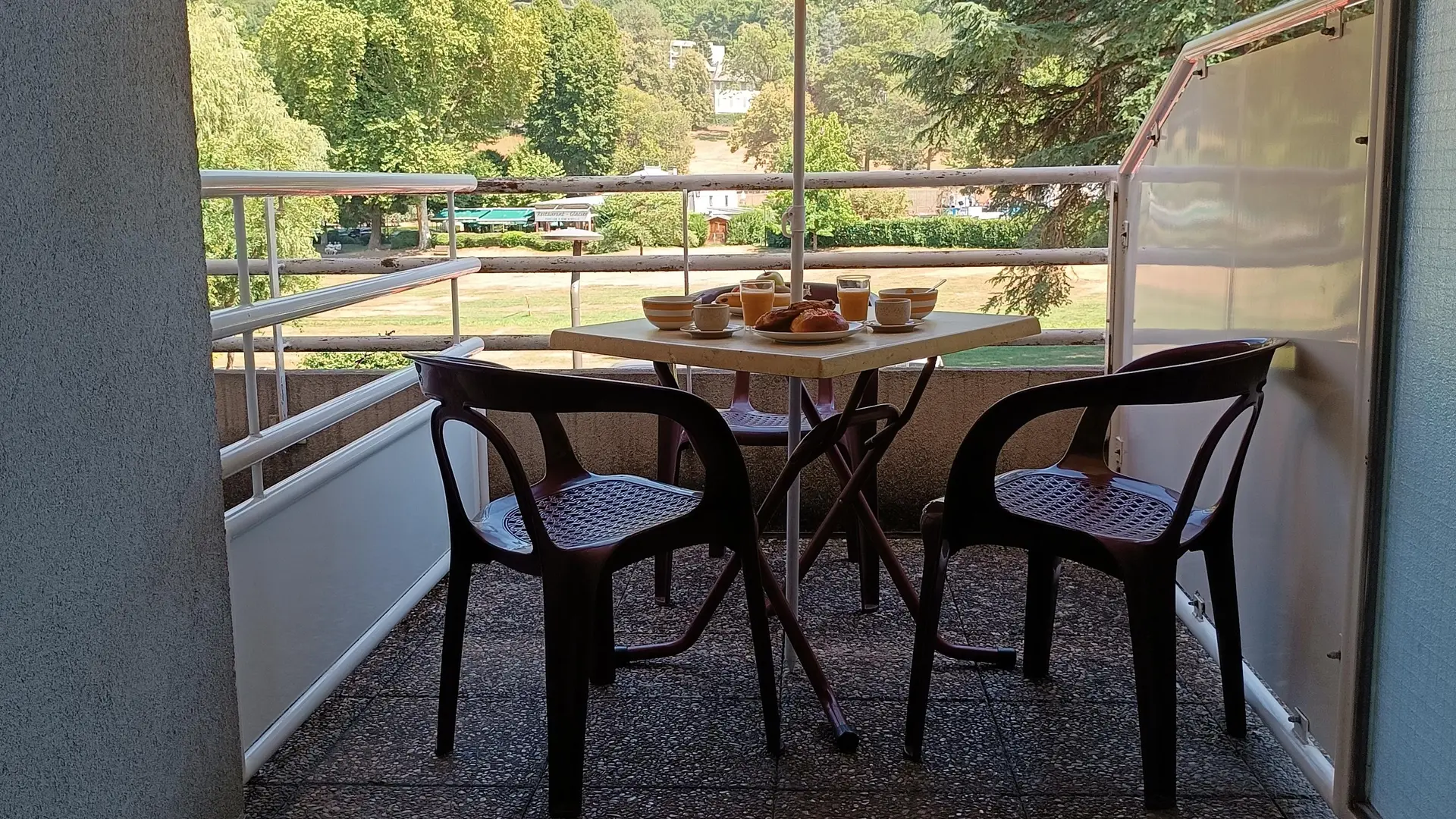 La terrasse est équipée d'une table et de deux chaises en plastique, sous un parasol. Un petit-déjeuner est servi sur la table et on aperçoit le parc d’Uriage en arrière-plan.