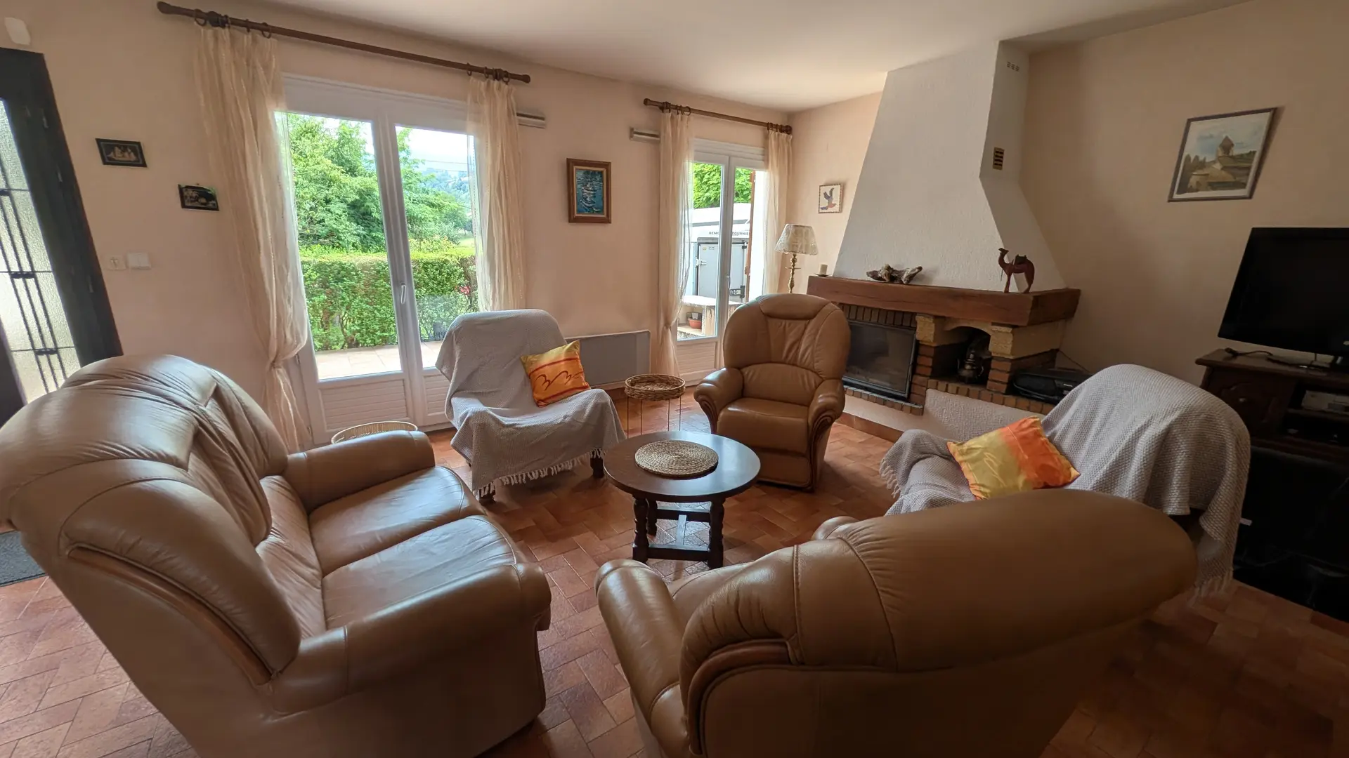 Un salon meublé de plusieurs canapés et fauteuils de styles variés, disposés autour d'une table basse ronde, fait face à une cheminée d'angle en pierre et brique. Des portes-fenêtres vitrées sur le mur du fond laissent voir de la verdure à l'extérieur, et un téléviseur est placé dans un meuble à droite.