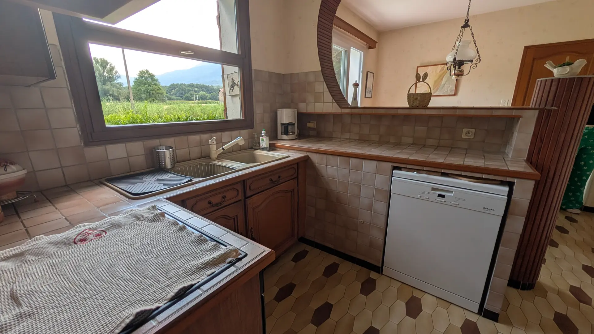 Une cuisine équipée, avec des meubles bas, un plan de travail comprenant un évier double face à une grande fenêtre, une cuisinière au premier plan à gauche et un lave-vaisselle blanc à droite.