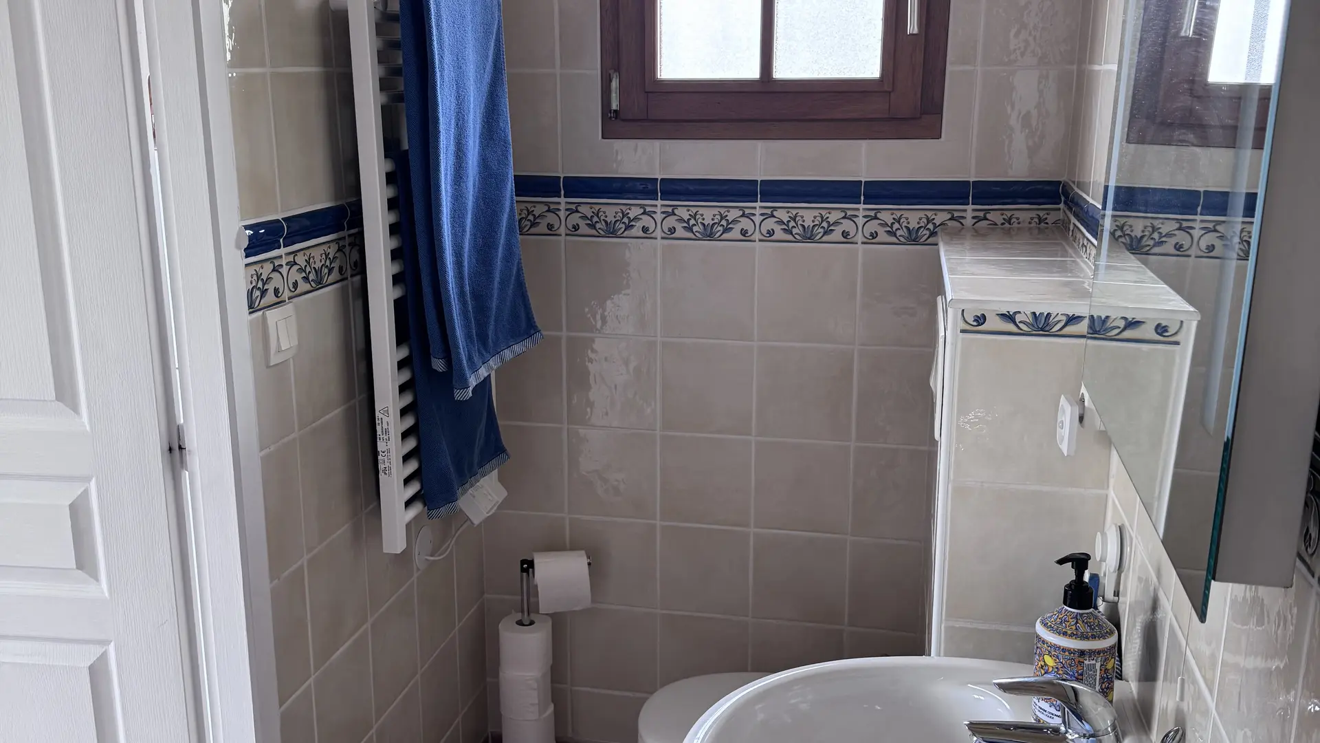 Cette salle d'eau entièrement carrelée de beige avec une frise décorative bleue comprend un lavabo colonne blanc au premier plan, un radiateur sèche-serviette mural où pendent deux serviettes bleues, et des toilettes installées au fond sous une petite fenêtre.