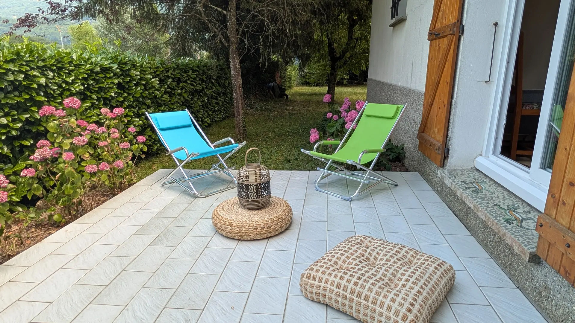 Une terrasse extérieure dallée, attenante à une maison et bordée par un jardin, sur laquelle se trouvent deux transats.