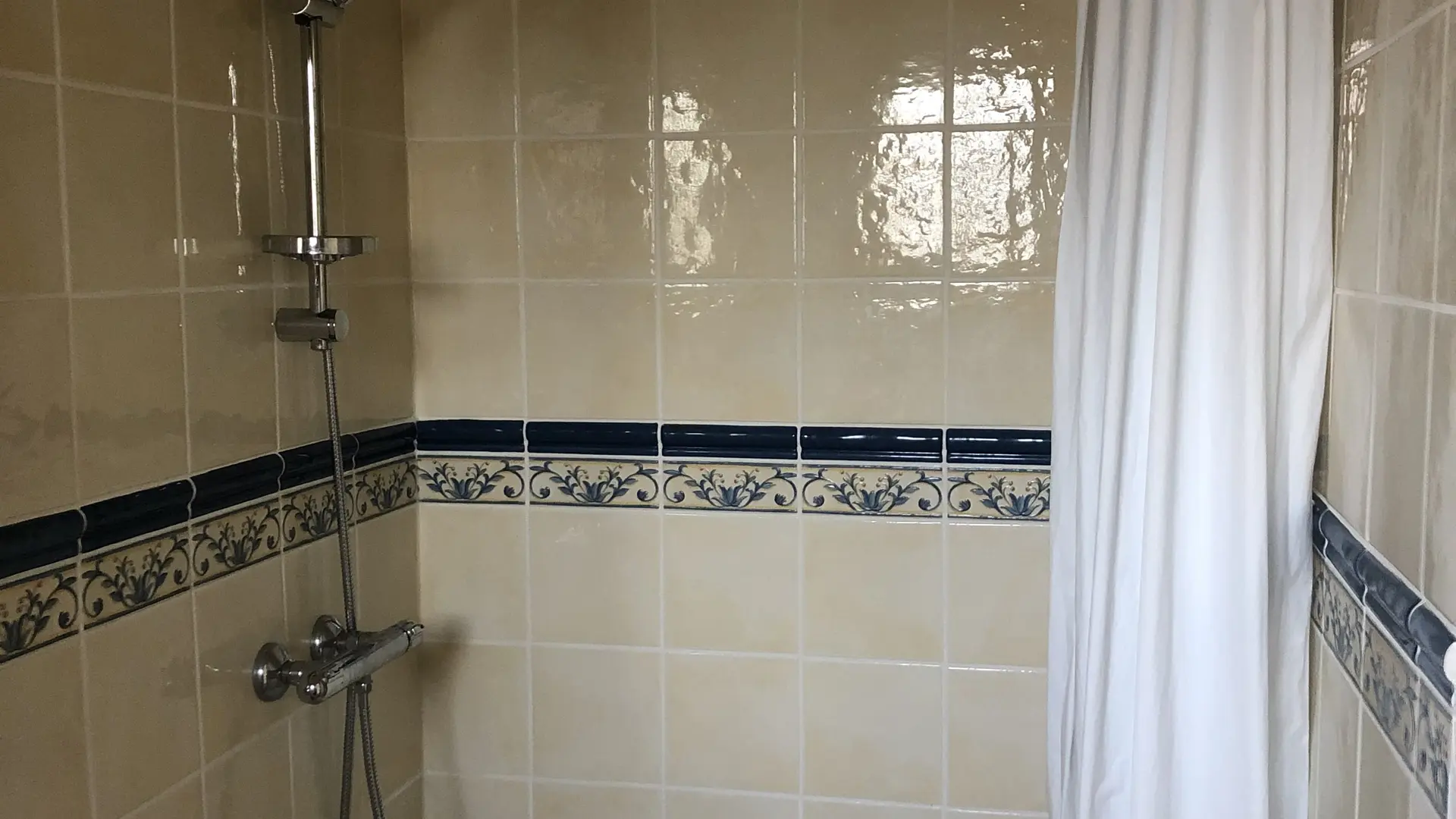 Un espace douche carrelé de beige avec la même frise décorative bleue et blanche, équipé d'une douchette et d'une tringle supportant un rideau de douche blanc.