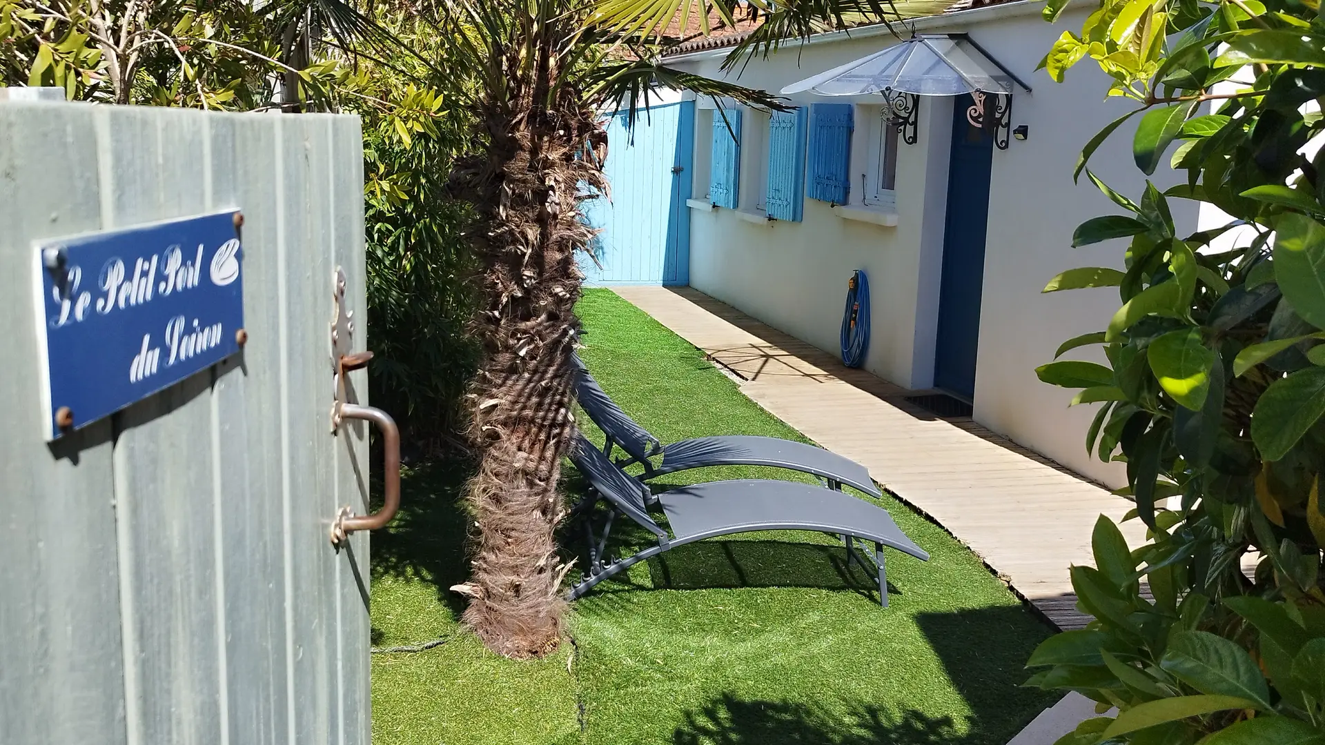 Portail d'entrée vers le jardin de la maison, avec deux bains de soleil, palmier.