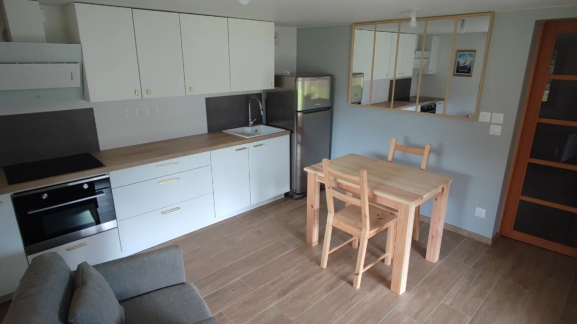 Cette pièce de vie comprend une cuisine ouverte avec un plan de travail blanc sur la gauche et un coin repas composé d'une table en bois et de chaises au centre.