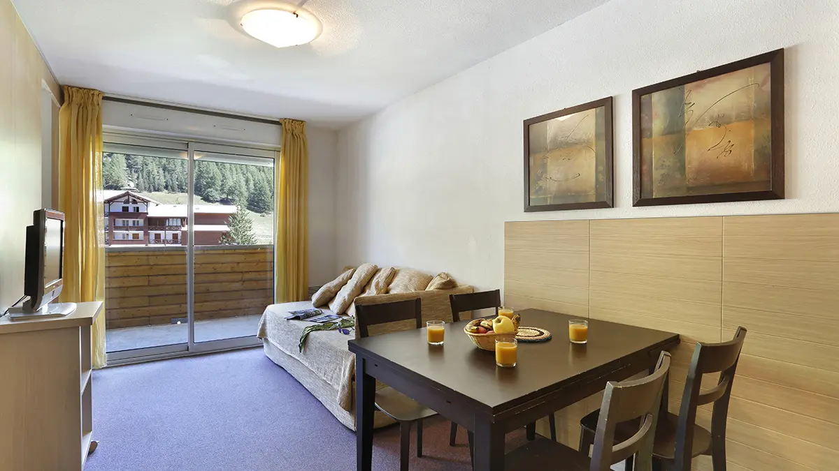 Séjour lumineux d'appartement avec canapé, meuble TV, TV, tabl à manger, porte-fenêtre donnant sur un balcon, vue sur la station