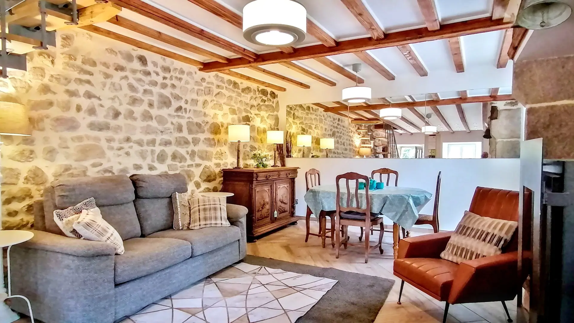 Une pièce de vie comprenant un coin salle à manger et un salon autour d'un poele à bois entouré d'un fauteuil et en face un canapé. La salle à manger comporte un buffet. Divers luminaires offrent un éclairage direct et indirect. Un miroir sur le mur du fond reflète la lumière de la pièce. On voit à droite l'escalier accédant aux chambres.