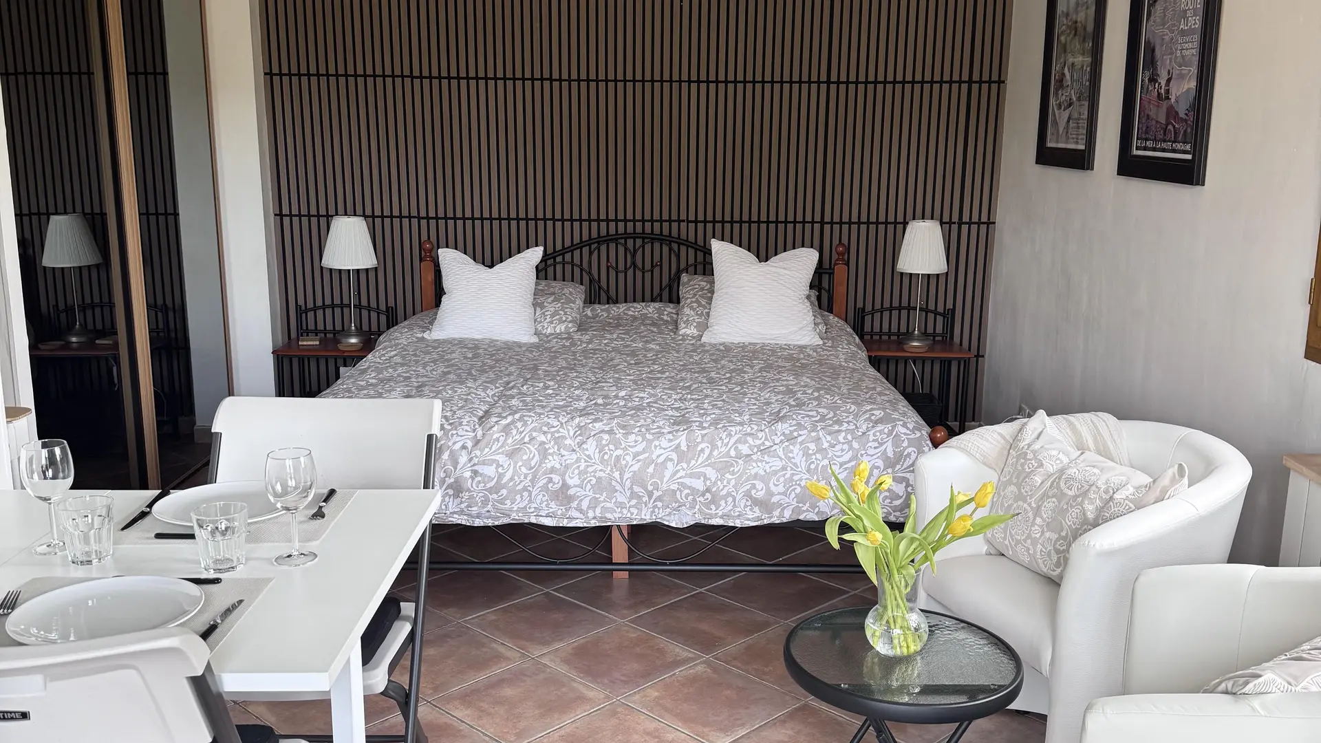 Cette chambre d'un studio moderne comprend un grand lit double fait de draps blancs adossé à une tête de lit marron, avec une petite table à manger à gauche et deux fauteuils blancs à droite entourant une table basse.