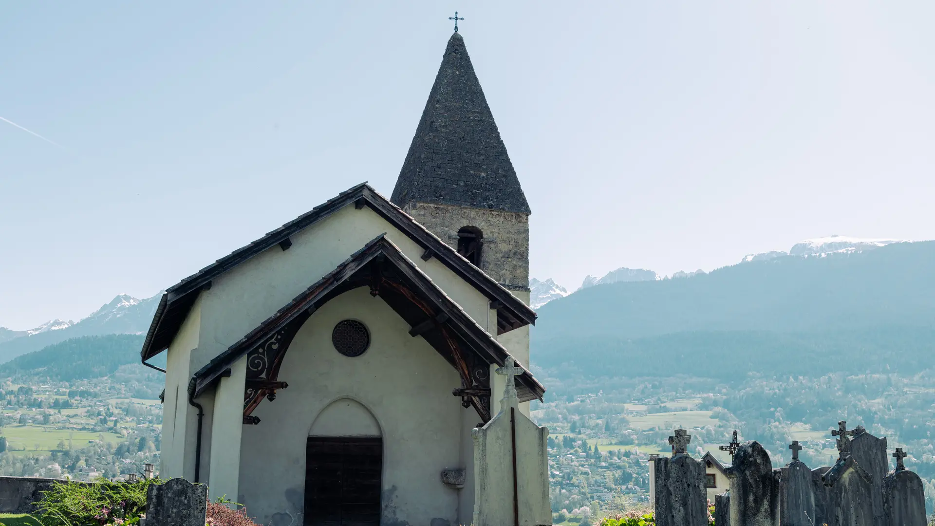 L'image montre la chapelle de Villeneuve d'Uriage, une structure en pierre claire avec un clocher pointu et un toit sombre, dressée sur une pente herbeuse où se trouvent des pierres tombales, avec un arrière-plan de montagnes et un ciel clair.