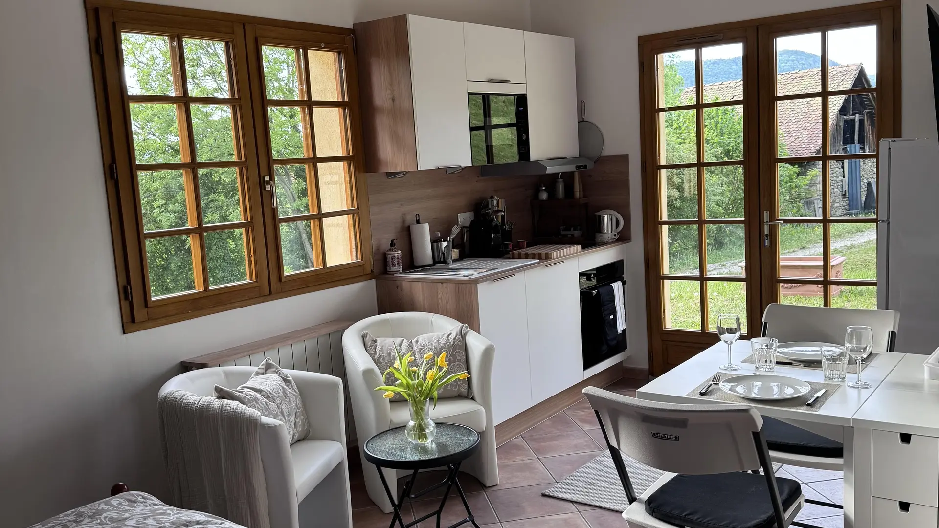 Ce studio lumineux réunit un coin nuit au premier plan, un espace cuisine aux meubles blancs et bois, deux fauteuils blancs autour d'une petite table basse fleurie, ainsi qu'une table à manger dressée Le studio ouvre sur l'extérieur par une double porte vitrée en bois.