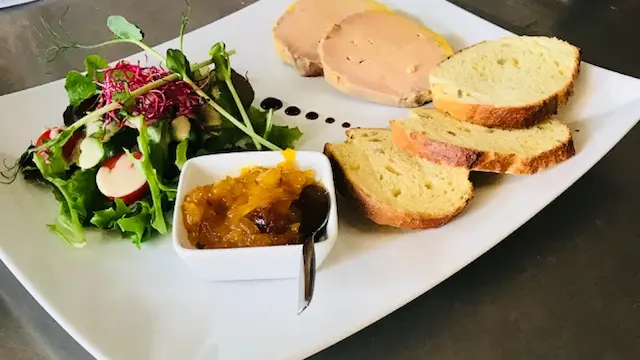 Nous avons une assiette de foie gras avec 2 médaillons de foie gras accompagnés de toast et d'une coupelle de compotée d'oignons et d'une farandole de salade. La décoration de l'assiette est agrémentée de points de vinaigre de Xérès.