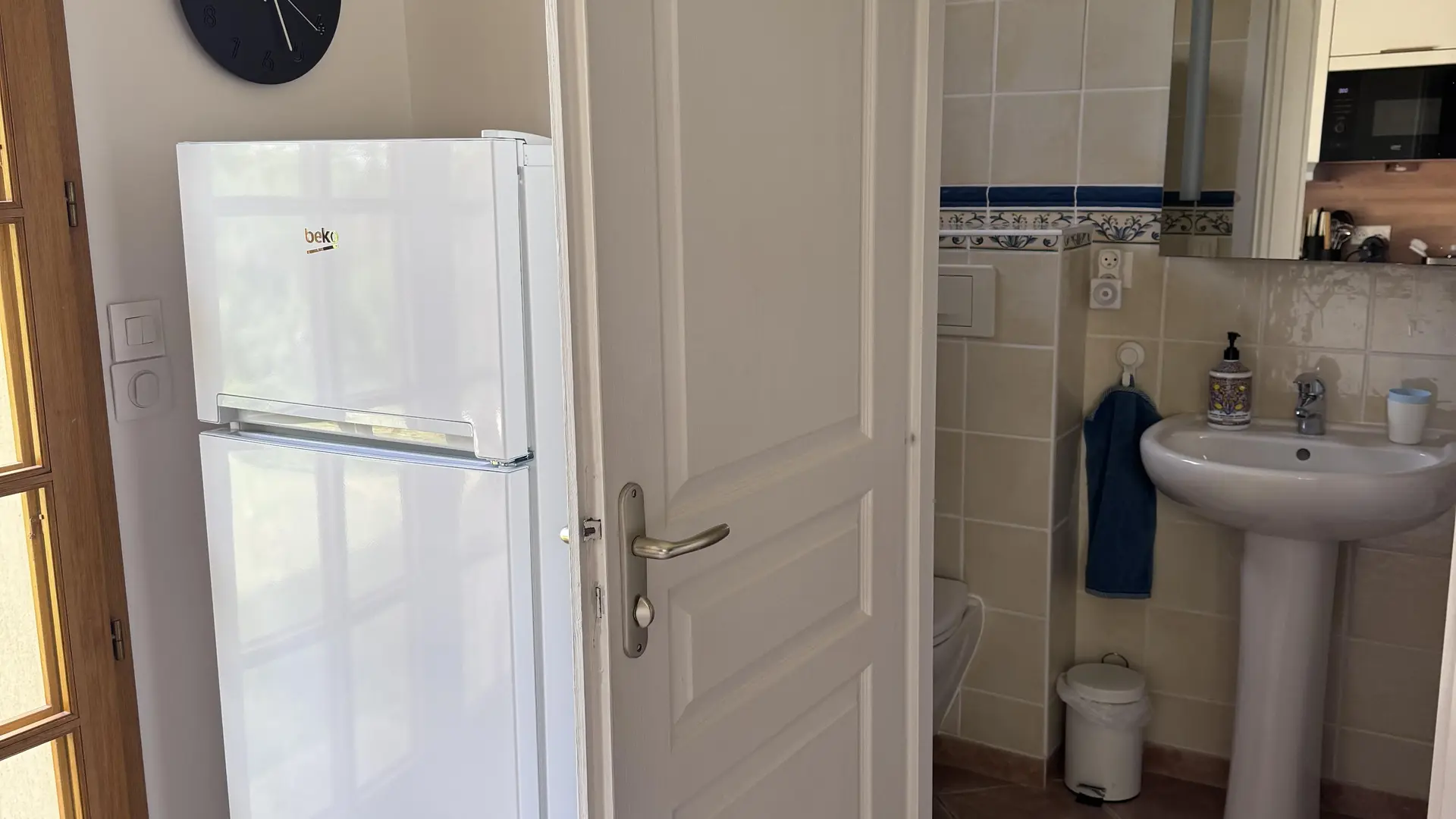 Cette vue montre une porte d'entrée blanche entrebâillée donnant sur une salle de bain avec un lavabo colonne, tandis qu'à gauche, un réfrigérateur blanc est installé à côté d'une porte vitrée en bois.