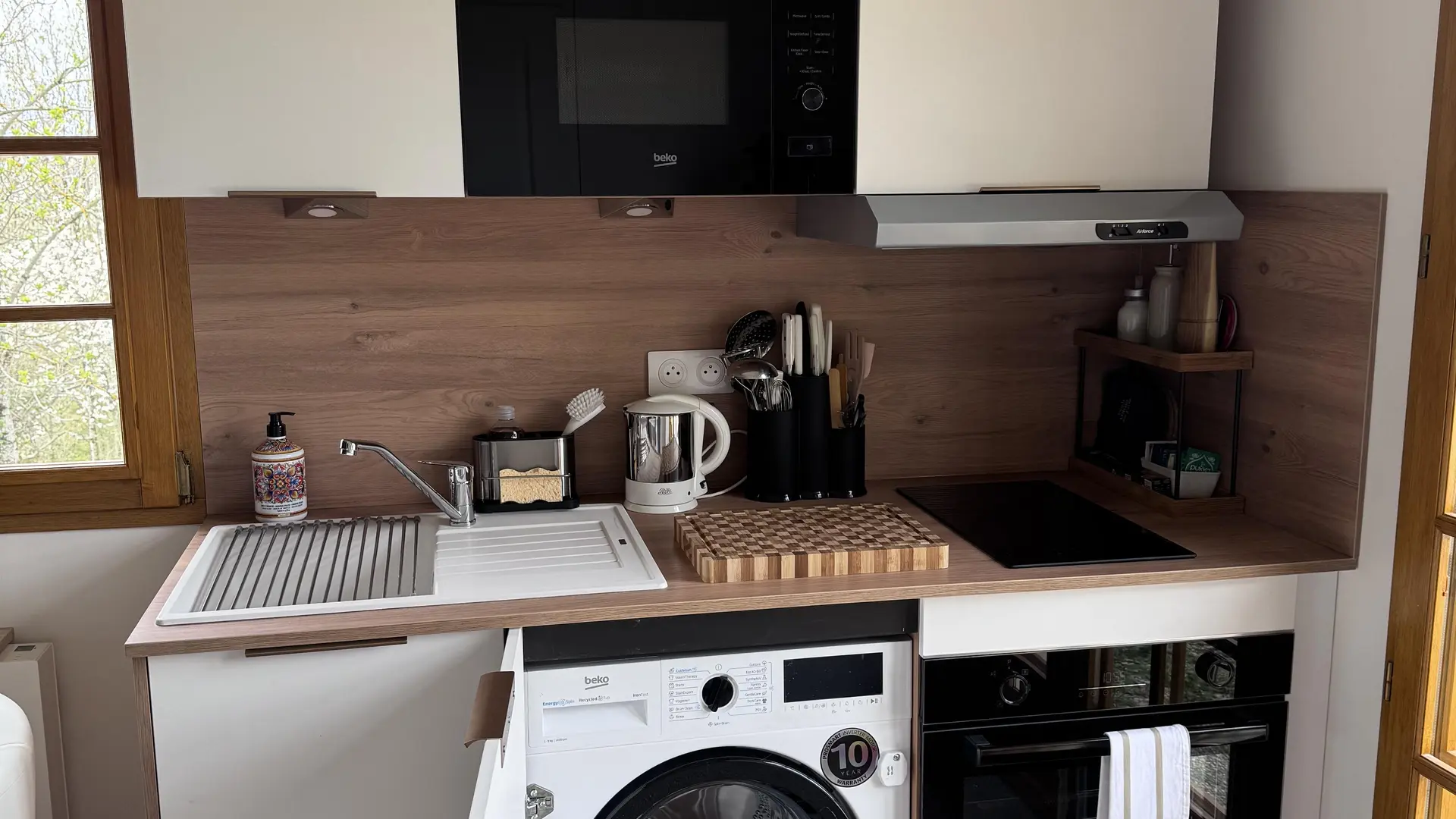 Cette cuisine moderne et compacte dispose d'un plan de travail avec un évier, de placards de rangement blancs suspendus, d'un four micro-ondes intégré et d'une petite fenêtre à deux battants en bois clair.