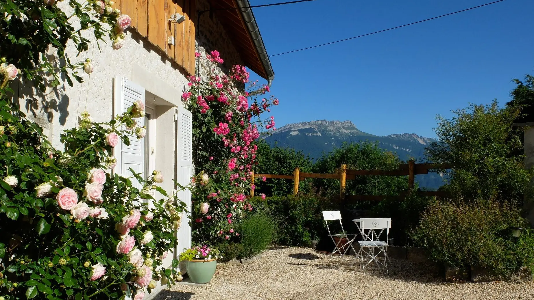 La façade d'une maison abritée baigne dans un soleil radieux. La porte d'entrée, cernée de rosiers, ouvre sur une allée où se trouvent une table et deux chaises positionnées près d'arbustes, orientées vers une montagne et son ciel bleu.