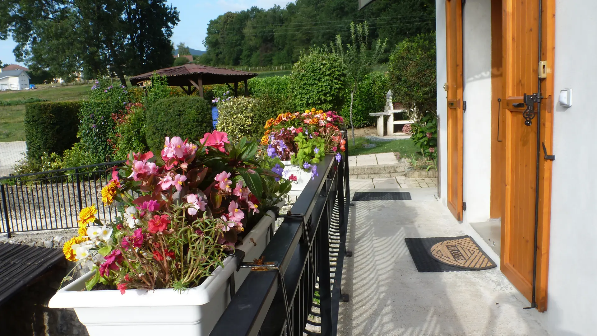 L'extérieur présente une petite terrasse ensoleillée devant une façade blanche avec des volets en bois orangé, bordée d'une balustrade sombre et de jardinières remplies de fleurs roses et rouges, avec un jardin verdoyant en arrière-plan.