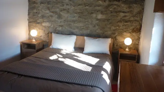Cette chambre présente un espace de repos avec un lit double agrémenté de deux tables de chevet surmontées de lampe. Le mur du fond est en pierre apparente.