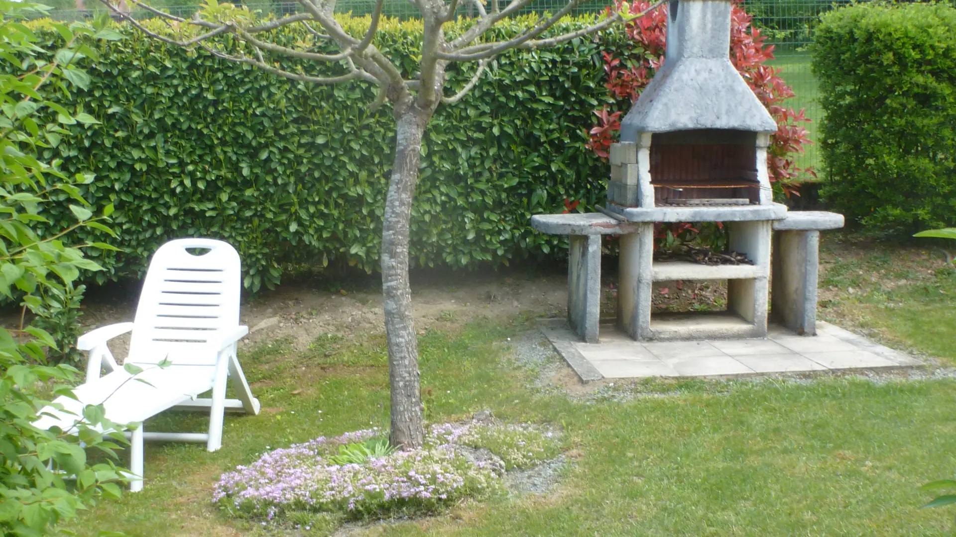 Le jardin est un espace extérieur clôturé par une haie, avec une zone de pelouse, un chemin pavé, un transat blanc et un barbecue en briques.