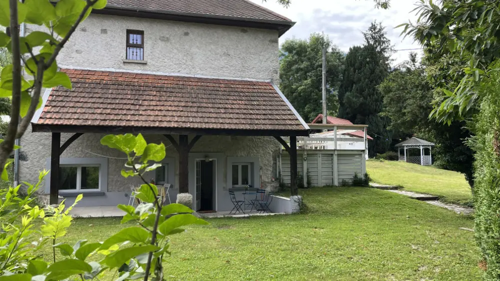 terrain est principalement herbeux et plat, avec des arbres et des arbustes dispersés. Une allée mène à la maison, et des bancs sont présents sous la partie couverte de la maison.