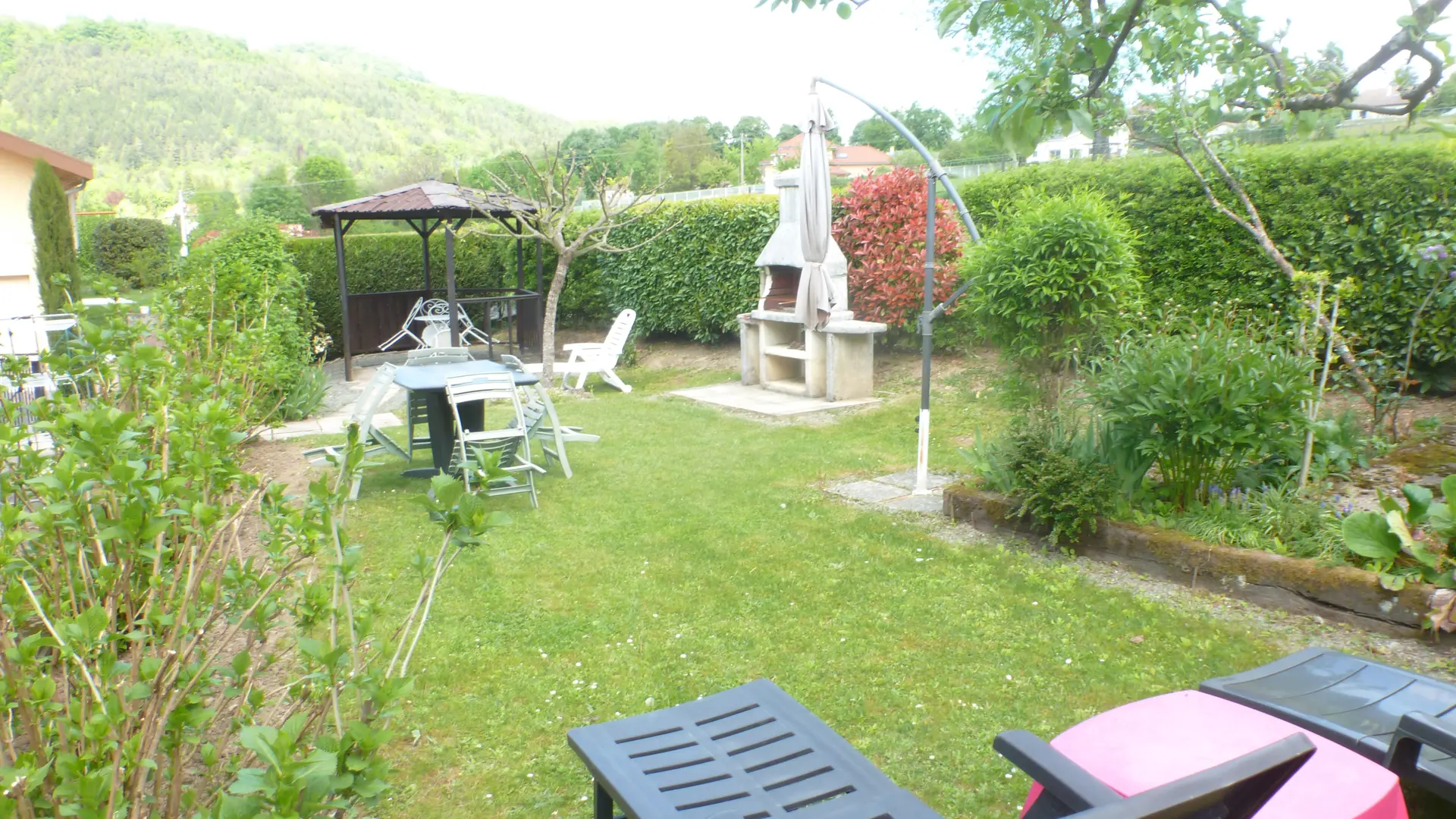 Ce jardin comprend une pelouse rectangulaire entourée de végétation et d'une haie, avec un barbecue fixe à l'arrière-plan, une table et des chaises au centre, et deux transats au premier plan.