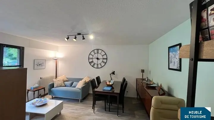 C'est une photo d'un salon-salle à manger, bien éclairé, meublé d'un canapé, d'une table basse, d'une table à manger et d'un buffet, le tout dans des tons neutres et avec un sol clair.