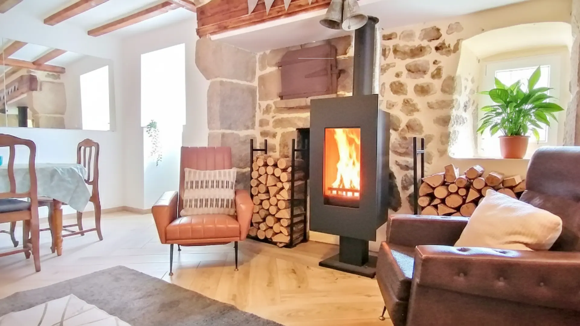 Un salon rustique et confortable avec un poêle à bois central et des murs en pierre apparente. Le sol est en parquet clair à chevrons.  Au centre, un poêle à bois moderne, noir et sur pied, est allumé et les flammes sont visibles derrière la vitre. Il est flanqué de piles de bûches de bois de chauffage. Le mur derrière le poêle est constitué de grandes pierres de taille.  Au premier plan à droite, un fauteuil club marron foncé avec un coussin blanc invite à la détente. Juste derrière lui, une fenêtre à carreaux en pierre est décorée d'une plante verte en pot. Au centre, un fauteuil en cuir brun-orange plus clair est tourné vers le poêle.  À l'arrière-plan à gauche, on aperçoit un coin repas avec une table recouverte d'une nappe claire et deux chaises en bois de style classique