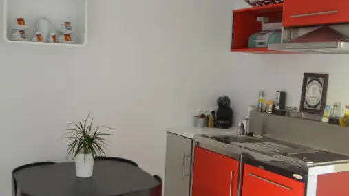 C'est une petite cuisine moderne et lumineuse avec des murs blancs, un sol carrelé blanc, des meubles de cuisine bas rouge vif et des meubles hauts rouges, ainsi qu'une table ronde noire avec quatre chaises assorties rangées dessous.