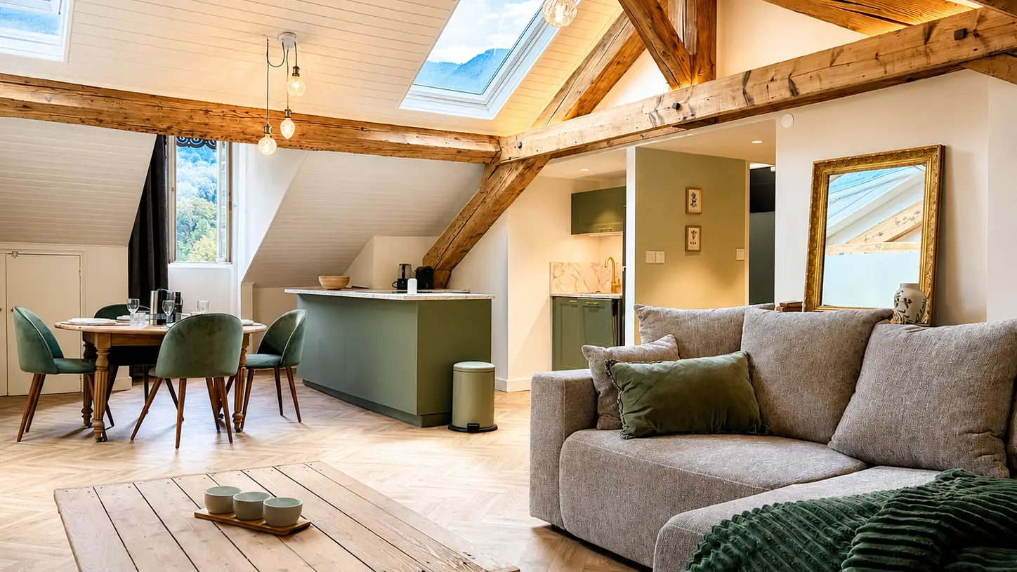 Cet appartement sous les toits offre un grand espace ouvert au sol en parquet, structuré par des poutres en bois et composé d'un salon avec un grand canapé d'angle en tissu, d'un îlot de cuisine vert et d'un coin repas sous des fenêtres de toit.