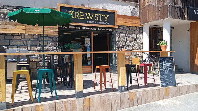 Vue de la terrasse extérieure en bois du restaurant, tabourets et chaises. Inscription Krewsty, street and fresh food'