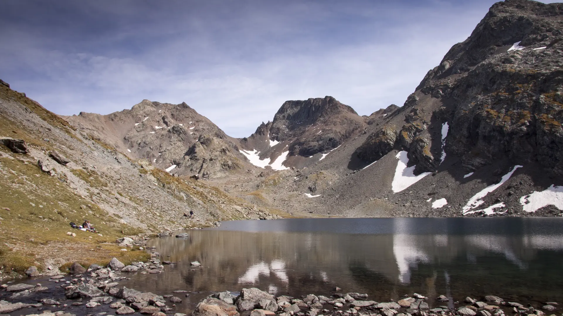 Le paysage présente un lac calme entouré de montagnes rocheuses et escarpées, avec quelques zones de neige résiduelle visibles sur les sommets.
